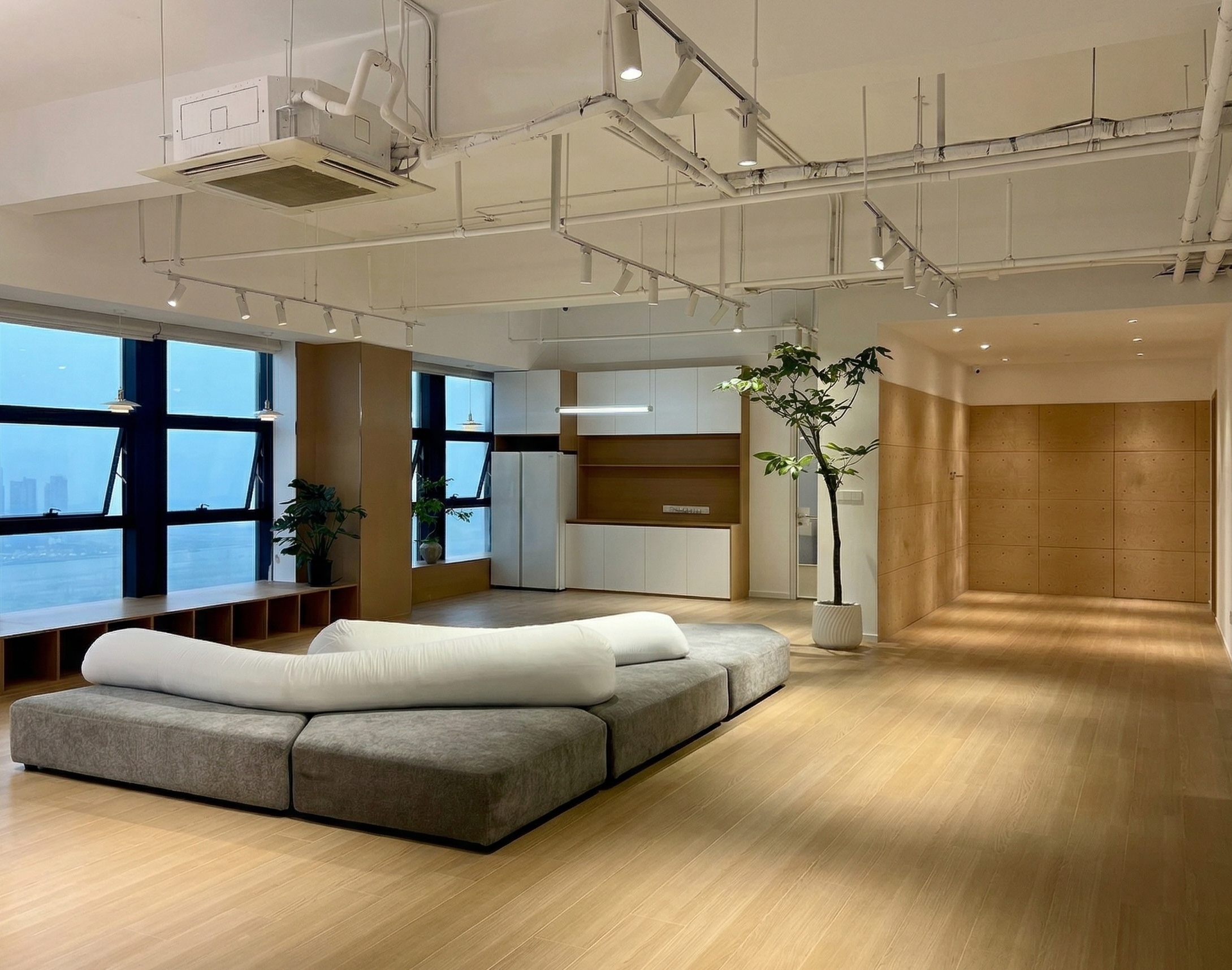 图片展示的是贾维斯联盟·杭州滨江“AI联合战队”场地的公区。画面中，左侧有大窗户，窗外景色隐约可见。窗边摆放着绿植，增添生机。地面铺设木质地板，显得温暖且有质感。左侧还有一排书架，书架旁是灰色的沙发，沙发上铺着白色靠垫。右侧是木质墙面，整体空间明亮且现代，与上下文介绍的“不止是办公室，更是‘AI超级实验室’”相契合，体现了空间的开放与舒适。