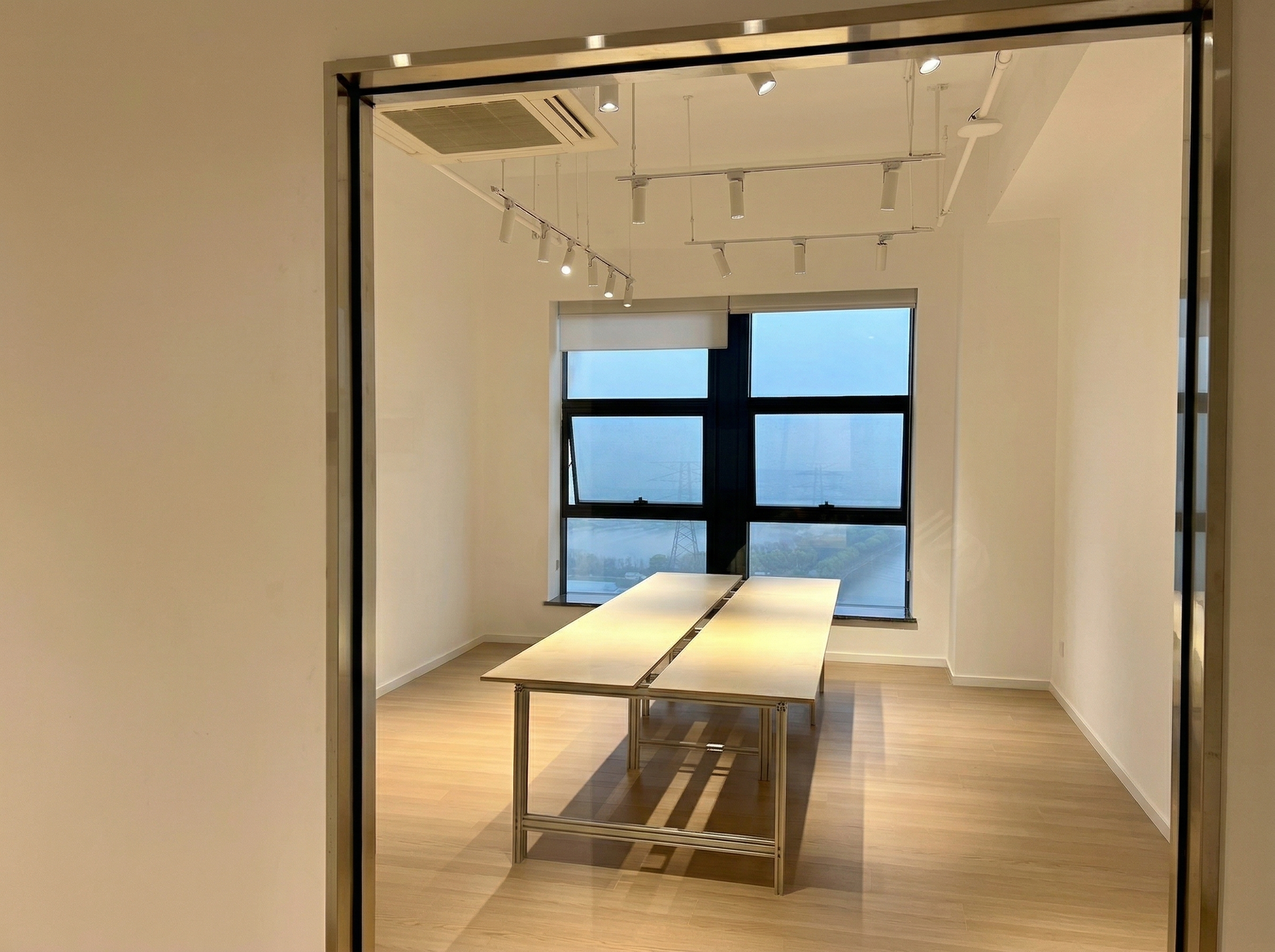 图片展示的是贾维斯联盟·杭州滨江“AI联合战队”场地内的一间会议室。房间内有两张并排的长桌，桌面为浅色，桌腿为金属材质。房间地面铺有木质地板，墙面为浅色。窗户较大，可看到窗外的建筑。房间顶部有多个嵌入式，可能是灯光或空调设备。此图与文档中介绍场地信息的内容相关，直观呈现了场地内会议室的环境。