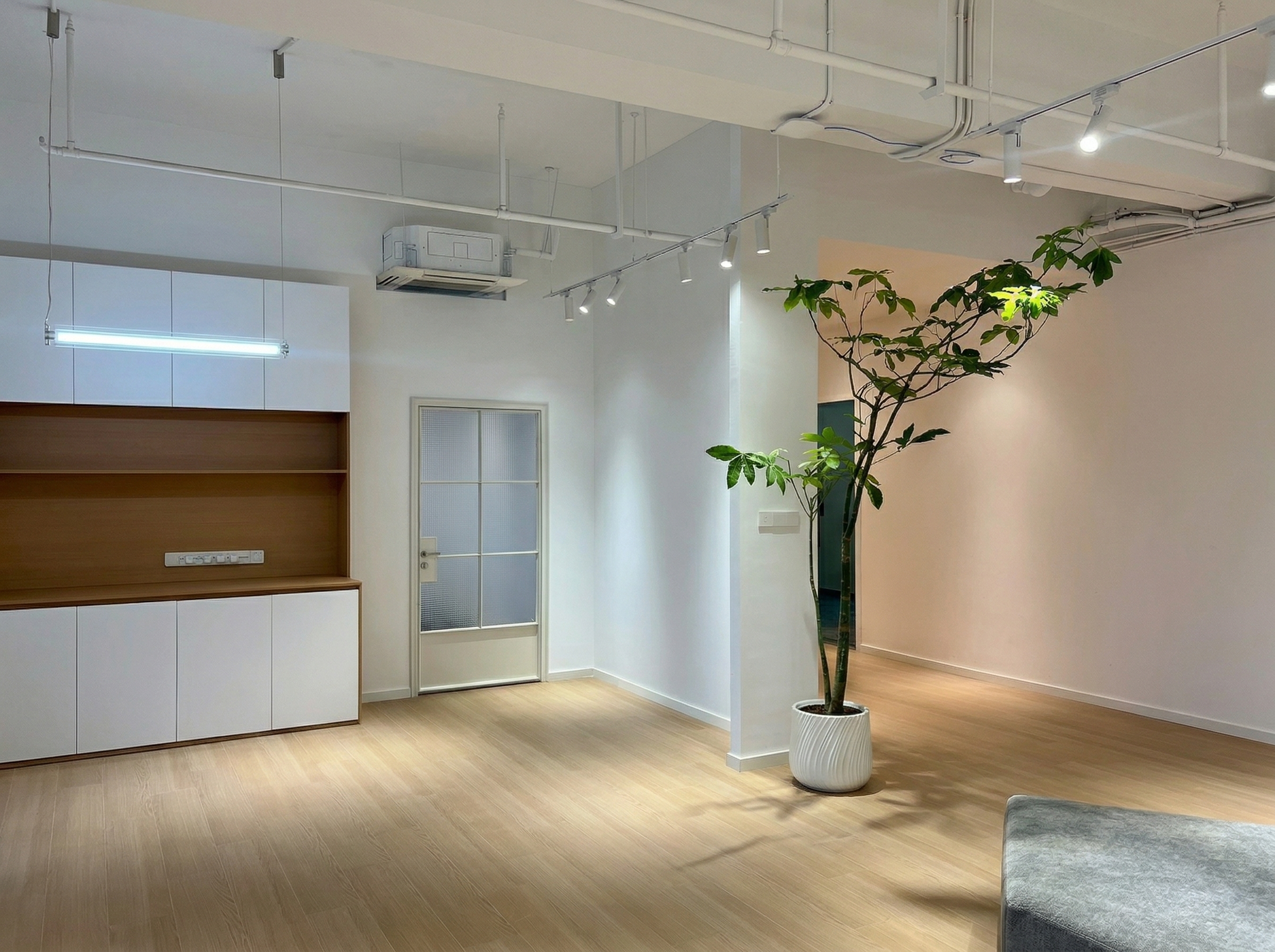 图片展示的是贾维斯联盟·杭州滨江“AI联合战队”场地的实拍场景。画面中，左侧有木质和白色柜体组合的储物区，上方装有空调。右侧是玻璃门，旁边放置着一盆高大的绿植。地面为木质地板，天花板上装有嵌入式灯具。该图片与文档中场地信息部分相关，直观呈现了场地的室内环境，与文档中不止是办公室，更是“AI超级实验室”的描述相呼应。