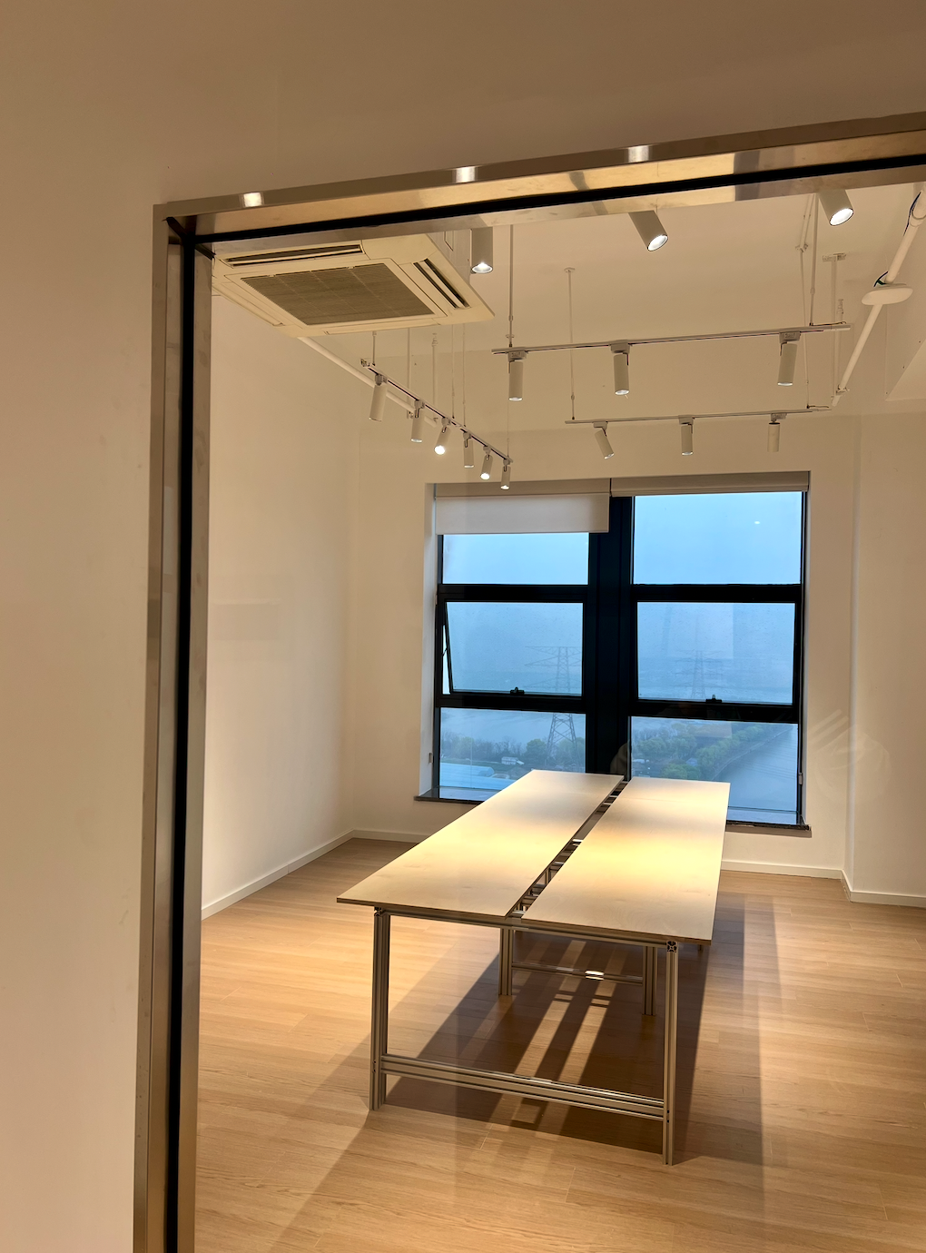 图片展示的是贾维斯联盟·杭州滨江“AI联合战队”场地内的一间空旷房间。房间内有一张长方形的白色桌子，桌面中间有黑色线条装饰，桌子下方有金属支架。房间地面铺有木质地板，墙面为白色。天花板上装有多个白色射灯 addCriterion图片内容