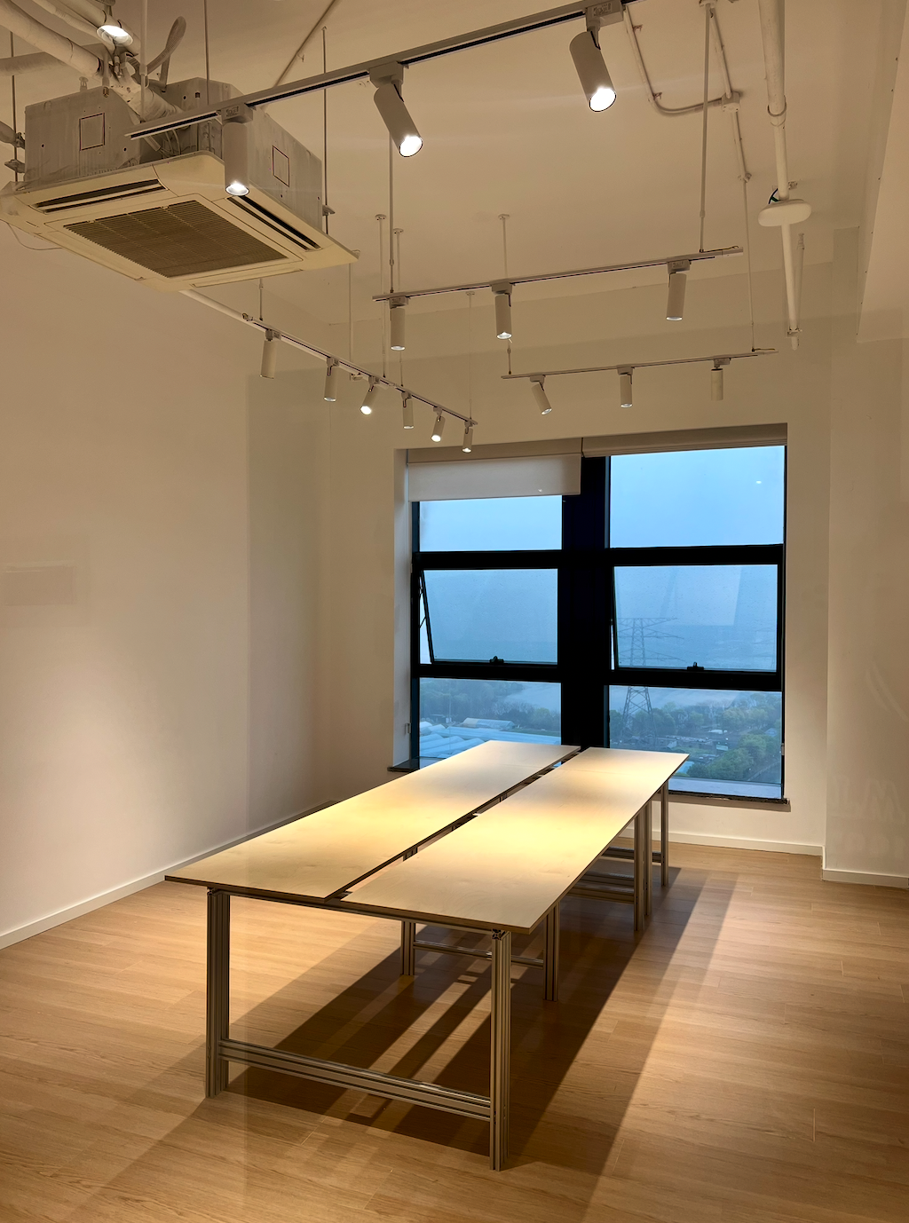 图片展示的是贾维斯联盟·杭州滨江“AI联合战队”场地内的一处公区。画面中，两排长桌并列摆放，桌面呈浅色，桌腿为金属材质。房间内有木质地板，天花板上安装有多个射灯和空调设备。背景是一扇大窗户，窗外景色隐约可见。该图片与文档中介绍场地信息的内容相关，直观呈现了场地内的部分环境。