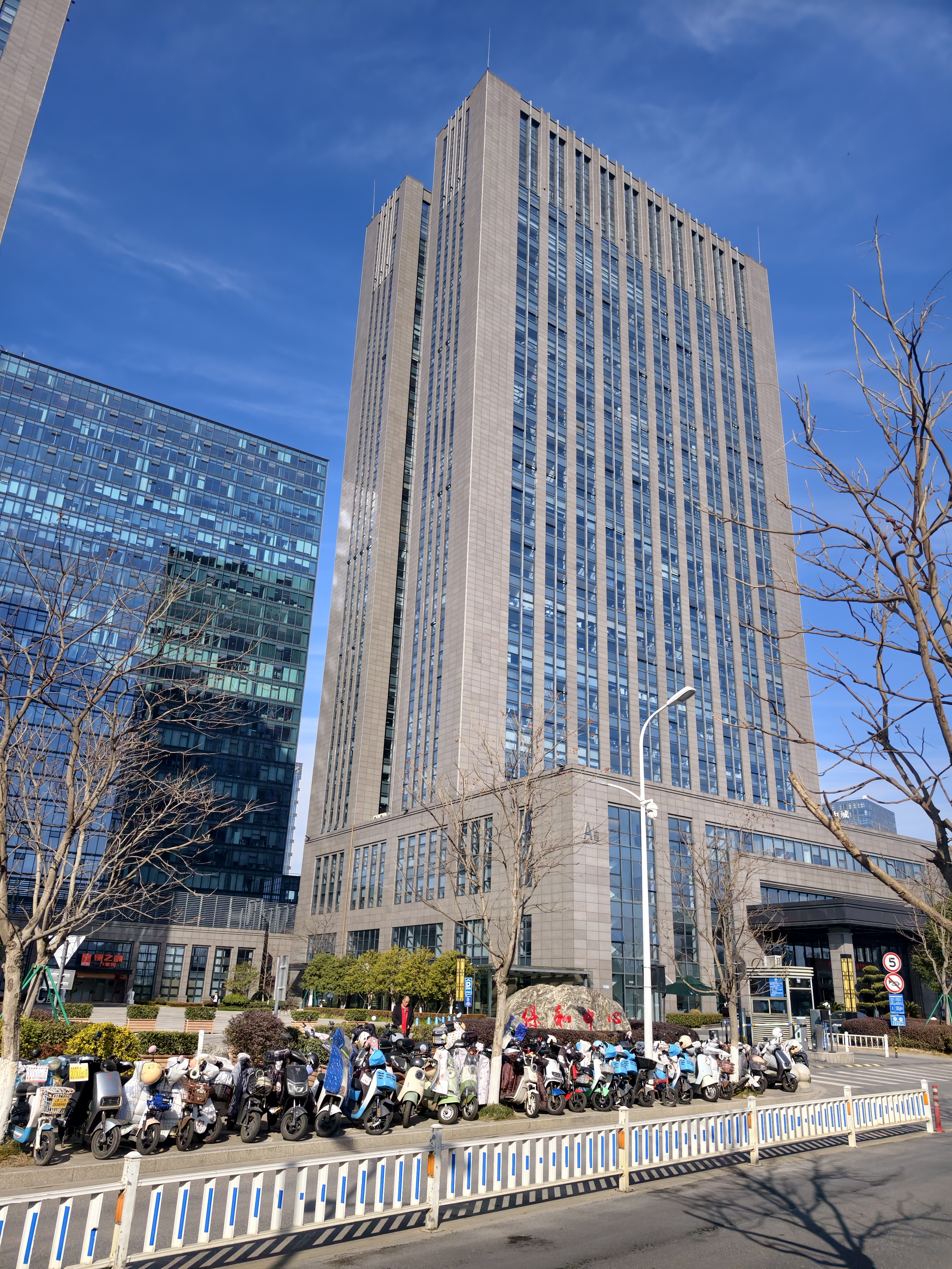 图片展示的是贾维斯联盟·杭州滨江“AI联合战队”场地外观。画面中，一座高耸的现代化玻璃幕墙建筑在蓝天白云下显得格外醒目，其前有整齐停放的电动车。建筑旁有几棵光秃秃的树木，地面上有蓝色和白色相间的护栏。建筑左侧还有一栋玻璃幕墙建筑。图片与文档中场地信息部分对应，直观呈现了场地外观，为读者提供视觉参考。